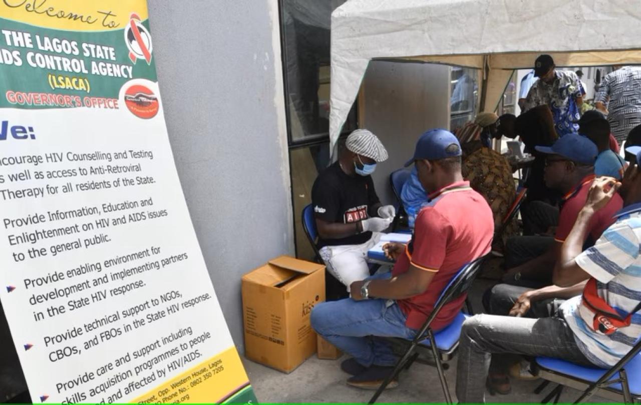 Patient wait in a street clinic beside a sign.