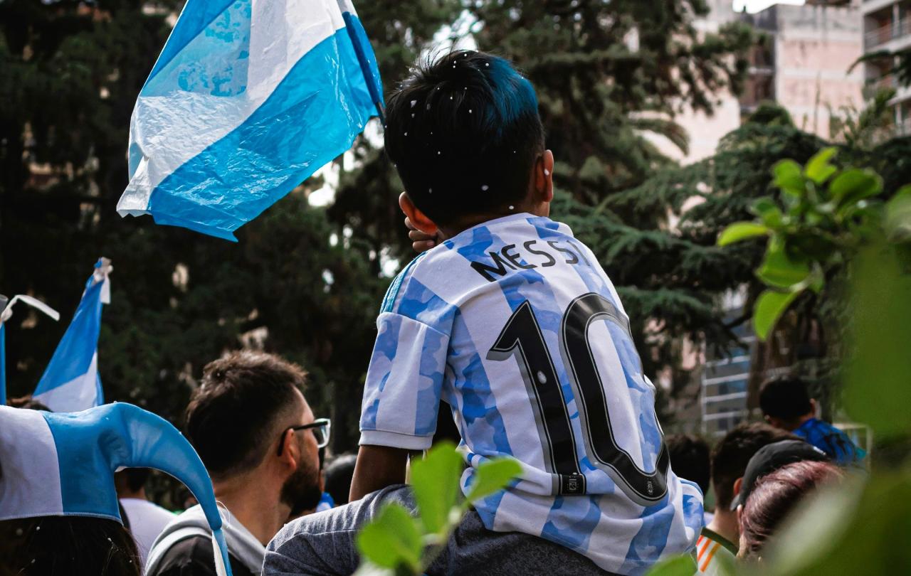 A child on the shoulders of a parent wears a light blue and white stripped football top, waves the Argentinian flag