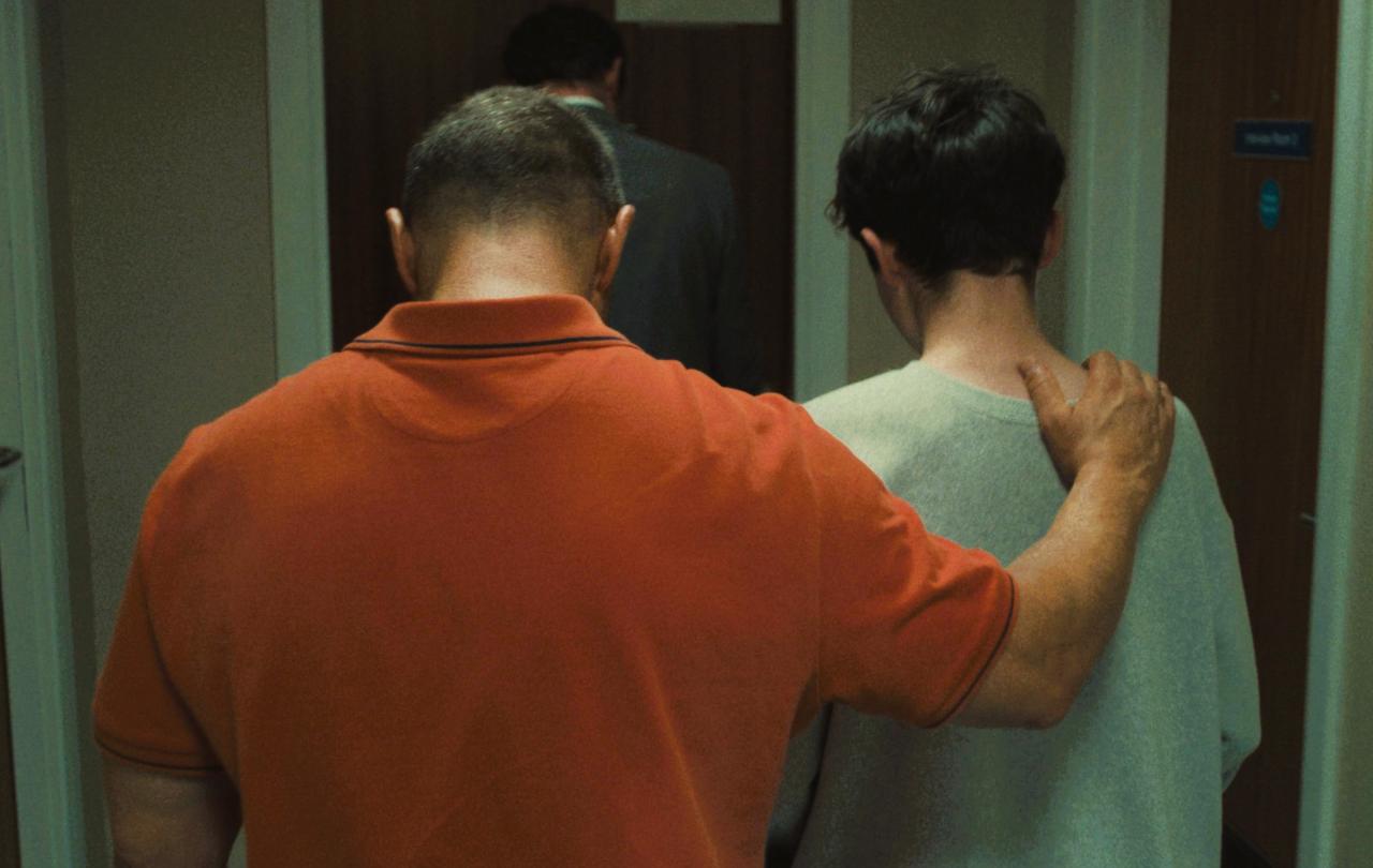 A father walks with his son away from the camera, his hand on his son's shoulder.