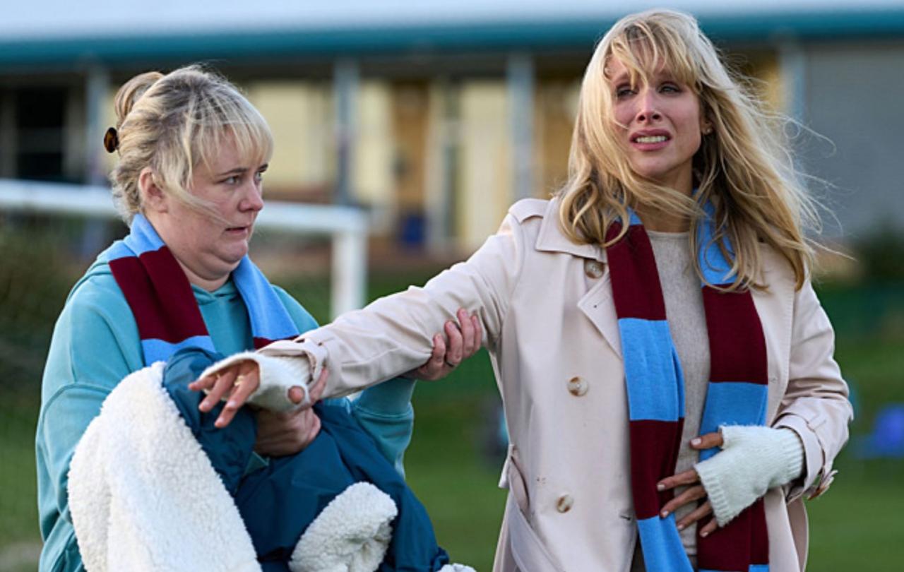 On the sidelines of a pitch a well-dressed mum hands a coat to a sceptical looking mum beside her.