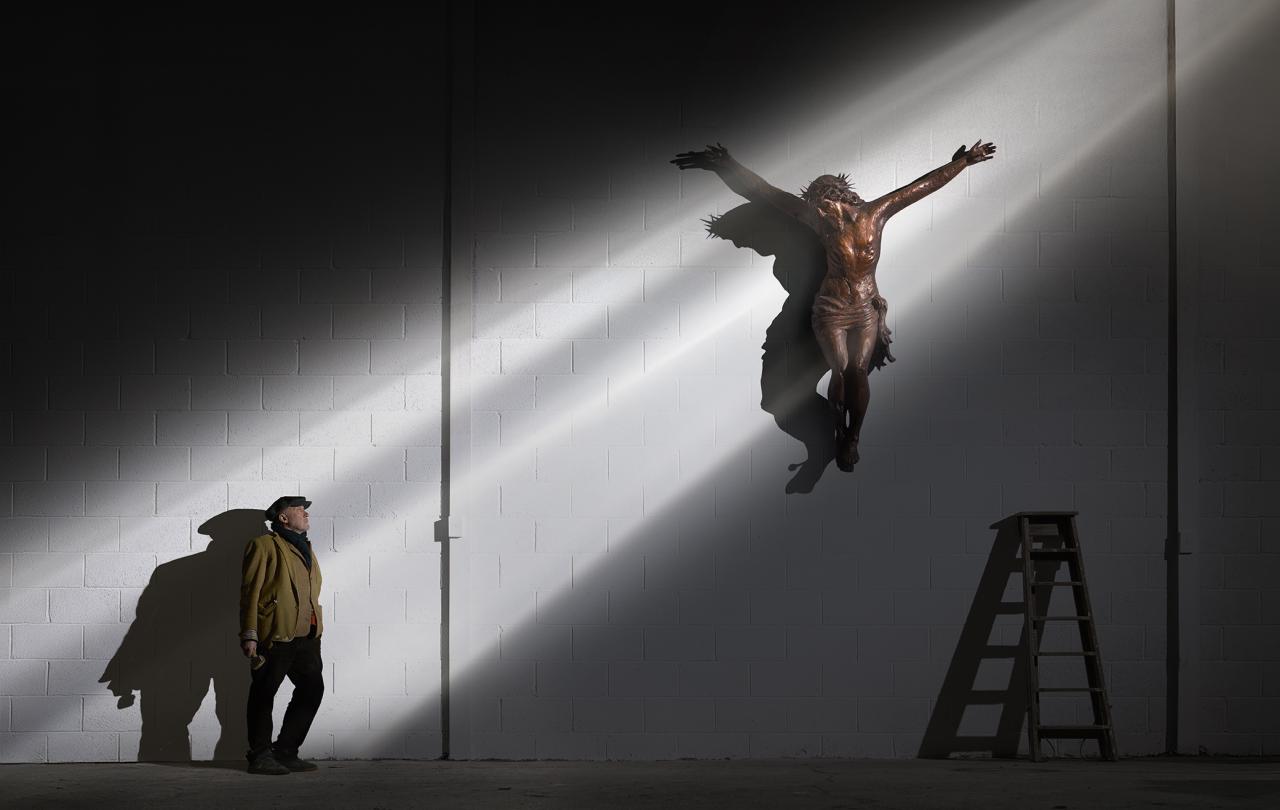 A man looks up a shaft of light that illuminates him and a crucifix higher up a wall.