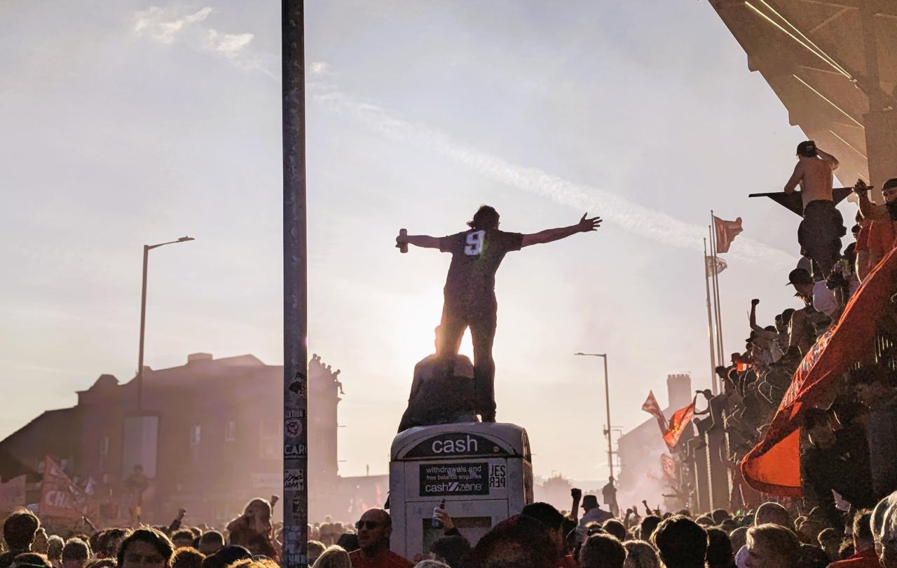 Amid celebrating football fans, one stands on top of a kiosk with outstretched arms.