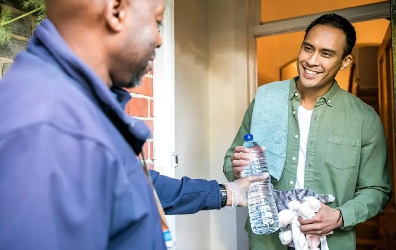 On a doorstep a man hands a bottle of water to another man.