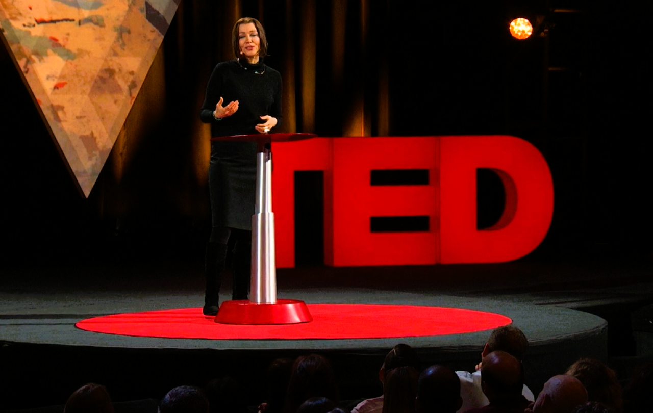 A speaker stands beside a podium in front of a TED sign.