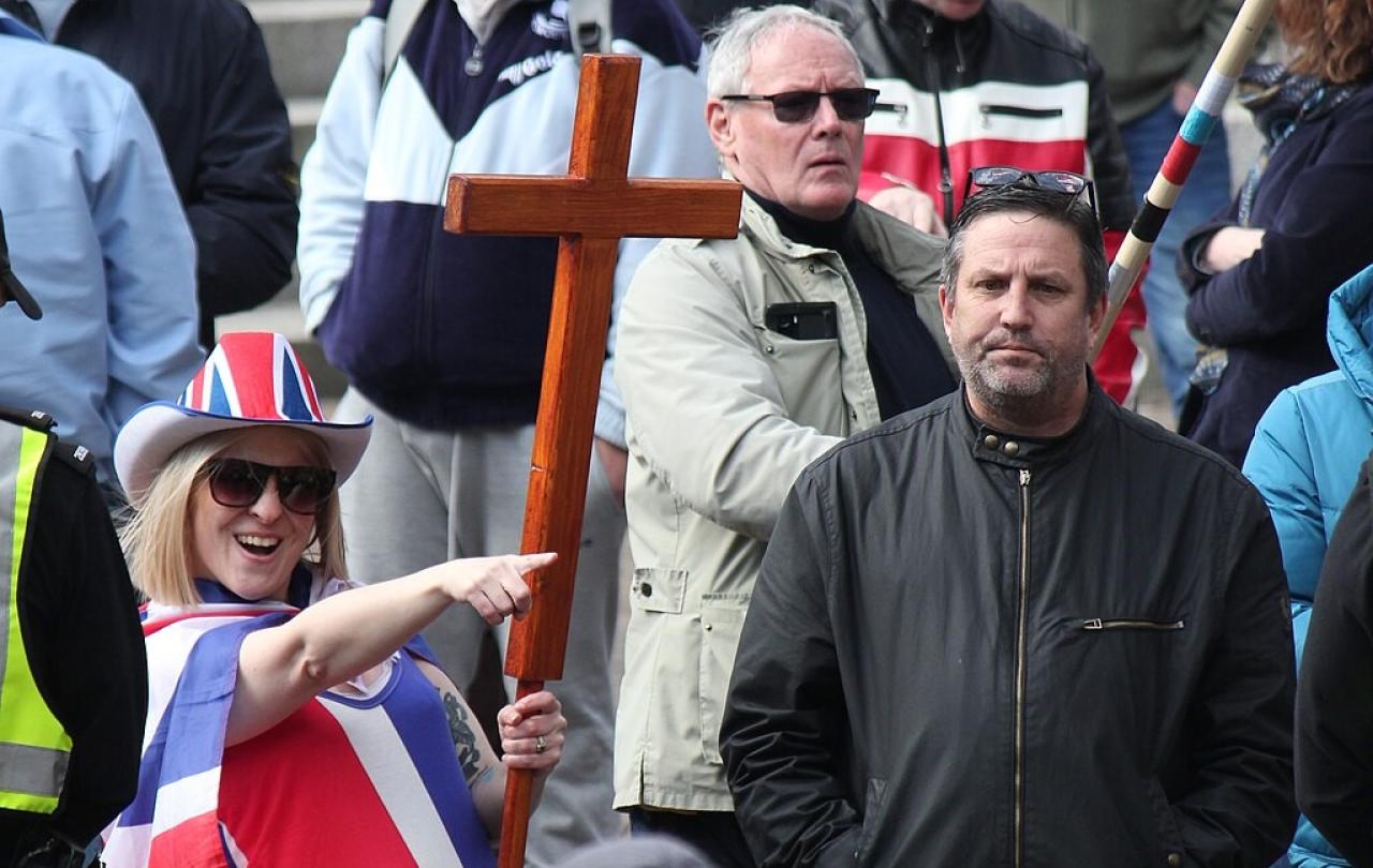 A protester wearing a Union Jack flag and hat and holding a cross, points while a man looks on.