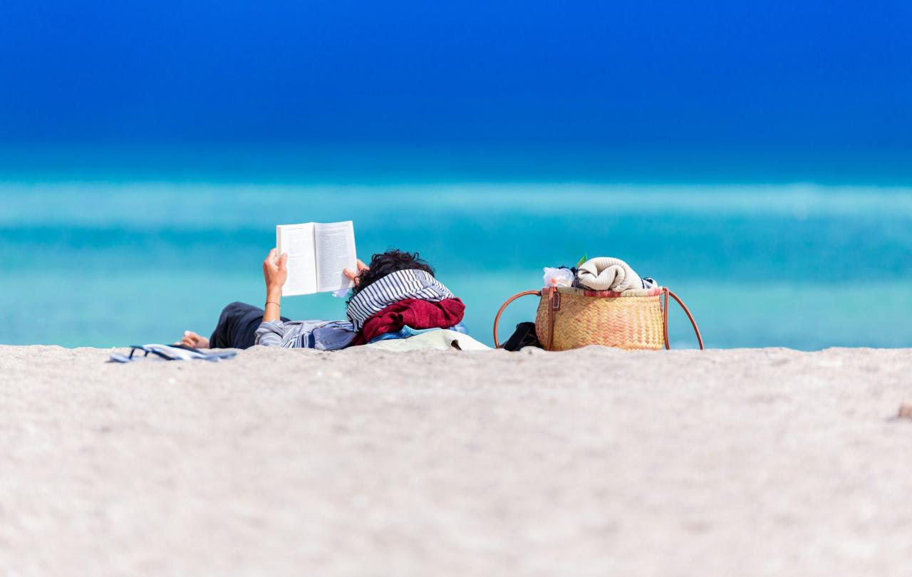 A person lying on a beach holds a book up to read.