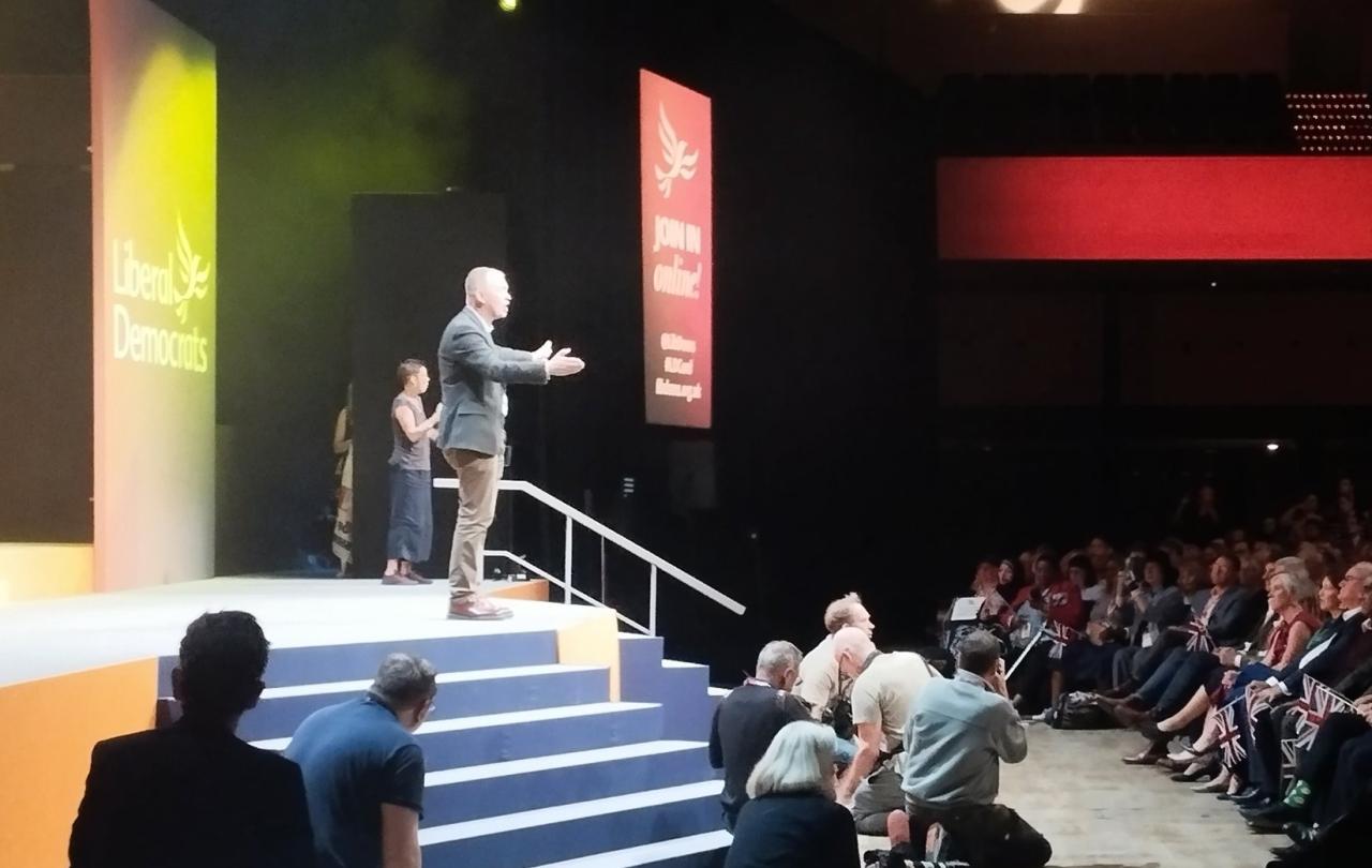 Tim Farron stands on a conference stage with arms wide