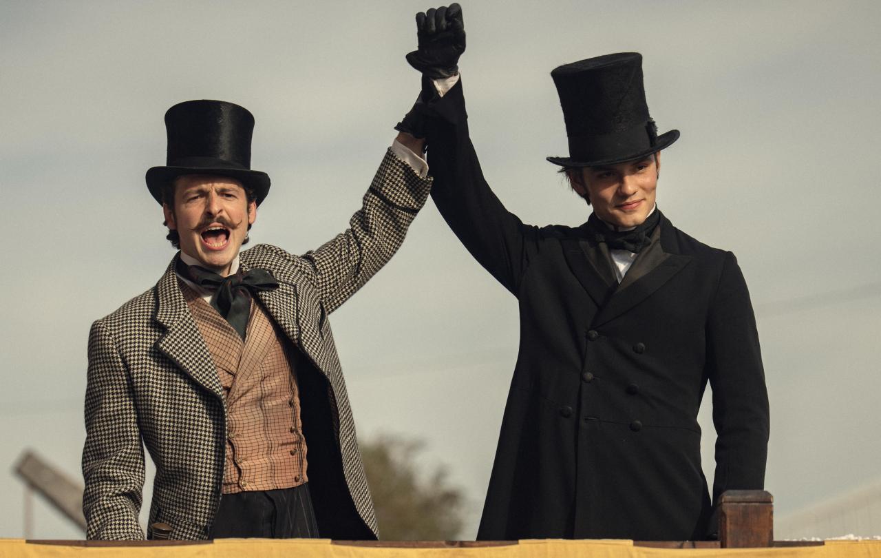 Dressed in Victorian clothes, two brothers raise their arms together.