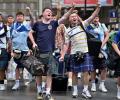 Scottish football fans wearing kilts march down a street singing and waving their arms alogt.