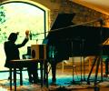 A pianist raises his arms while sitting at a grand piano amid recording equipment.