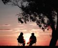 Under a tree, backlit by a sun set, two people sit in chairs outside and talk.