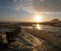 A rockpool on a beach reflects the sun, a castle stands beyond the sand dunes