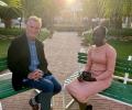 A TV interviewer sits across from a young woman in an outdoor setting