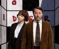 Two TV characters, a man and a woman, stand in front of a crossword cover walls of a room. 