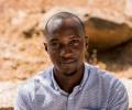 A Nigerian man looks up towards the camera, behind him is dusty ground