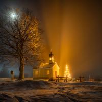 A nocturnal snow-covered scene of a tree, chapel and Christmas tree casting shadows.