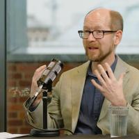 A podcast guest sits in front of a mic, gesturing with their hands.