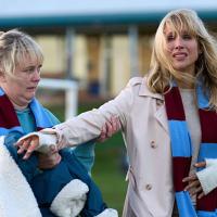 On the sidelines of a pitch a well-dressed mum hands a coat to a sceptical looking mum beside her.